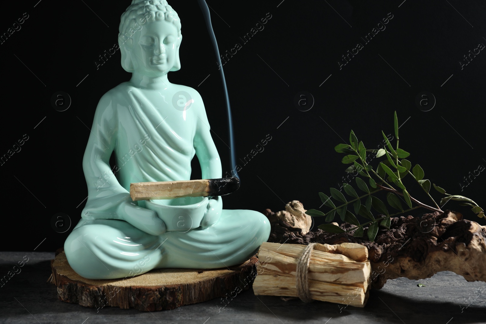 Photo of Smoldering palo santo stick, Buddha statue and snag on grey textured table against black background, closeup