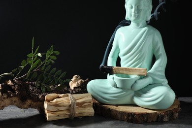 Photo of Smoldering palo santo stick, Buddha statue and snag on grey textured table against black background, closeup