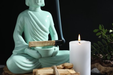 Photo of Smoldering palo santo stick, burning candle and Buddha statue on table against black background, closeup