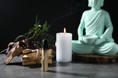 Photo of Smoldering palo santo stick, burning candle, Buddha statue and snag on grey textured table against black background, closeup