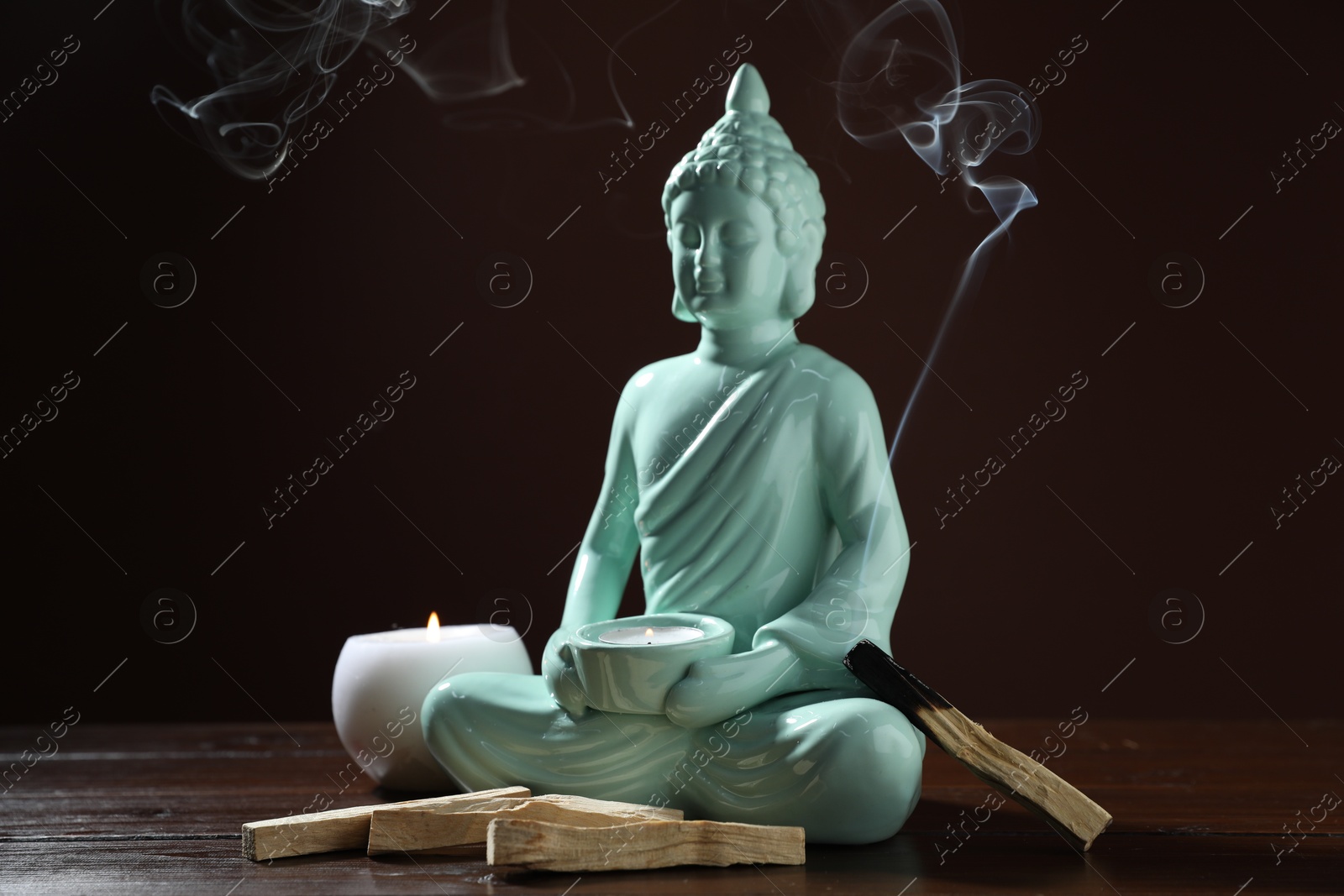 Photo of Smoldering palo santo stick, burning candles and Buddha statue on wooden table against dark background, closeup