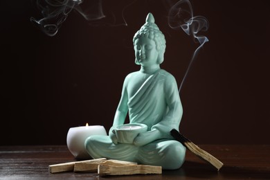 Photo of Smoldering palo santo stick, burning candles and Buddha statue on wooden table against dark background, closeup