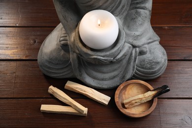 Photo of Palo santo sticks and statue with burning candle on wooden table, closeup