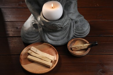 Photo of Palo santo sticks and statue with burning candle on wooden table, closeup