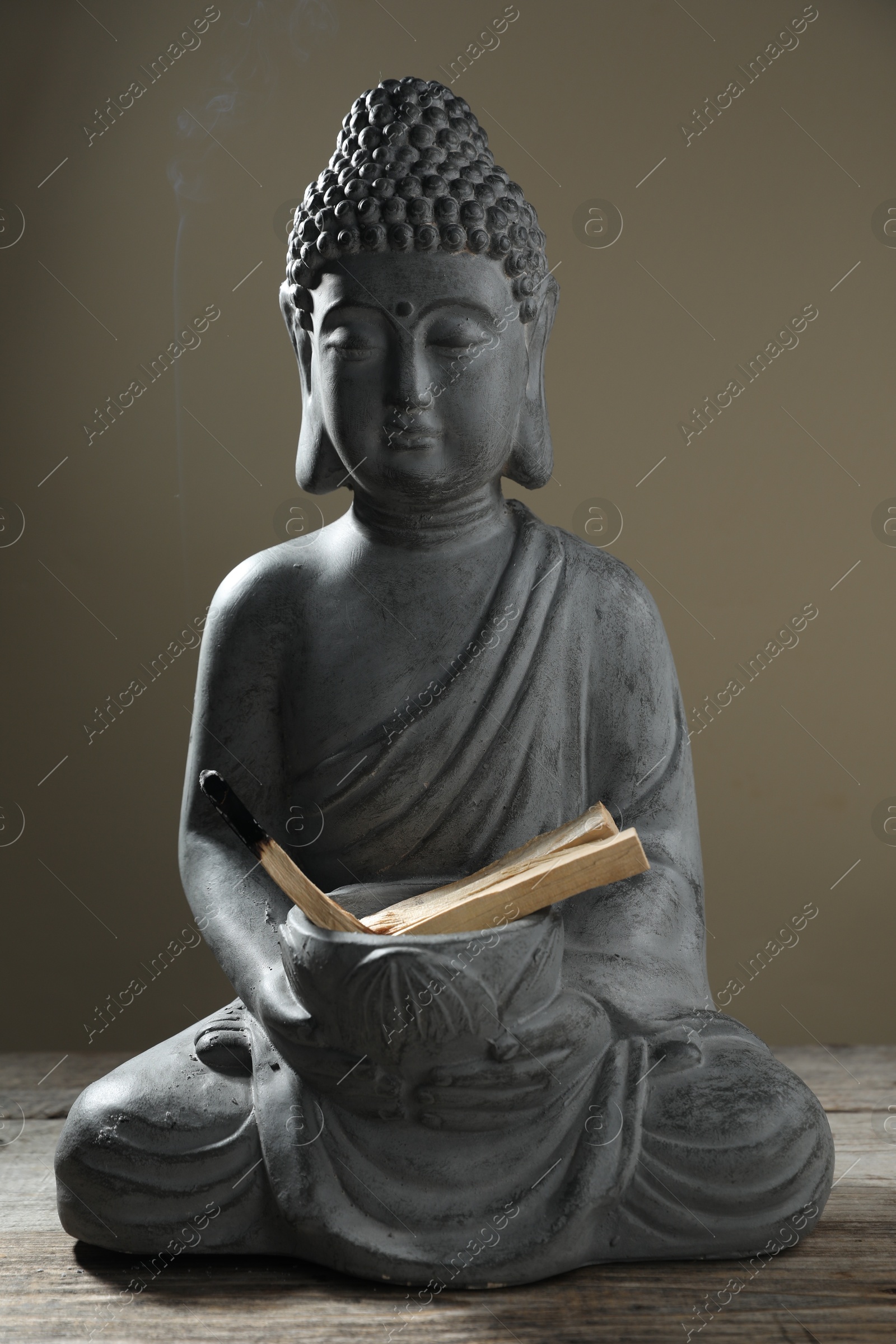 Photo of Smoldering palo santo stick and Buddha statue on wooden table against grey background, closeup