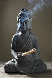 Photo of Smoldering palo santo stick and Buddha statue on wooden table against grey background, closeup