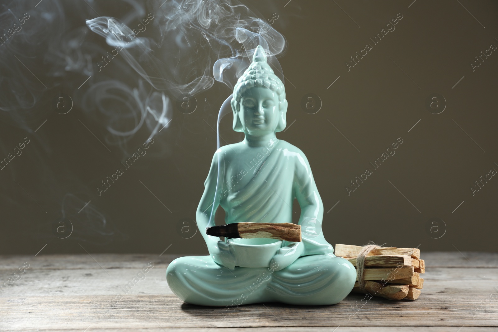 Photo of Smoldering palo santo stick and Buddha statue on wooden table against grey background