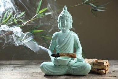 Photo of Smoldering palo santo stick, Buddha statue and beautiful branches on wooden table against grey background, closeup