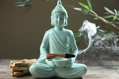 Photo of Smoldering palo santo stick, Buddha statue and beautiful branches on wooden table against grey background, closeup