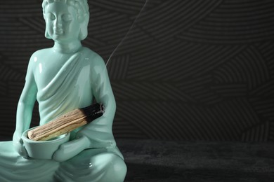 Photo of Smoldering palo santo stick and Buddha statue on grey textured table against black background, closeup. Space for text