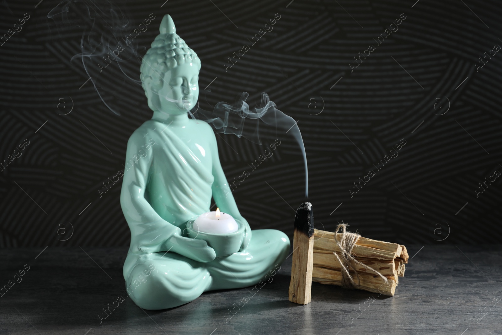 Photo of Smoldering palo santo stick and Buddha statue with burning candle on grey textured table against black background