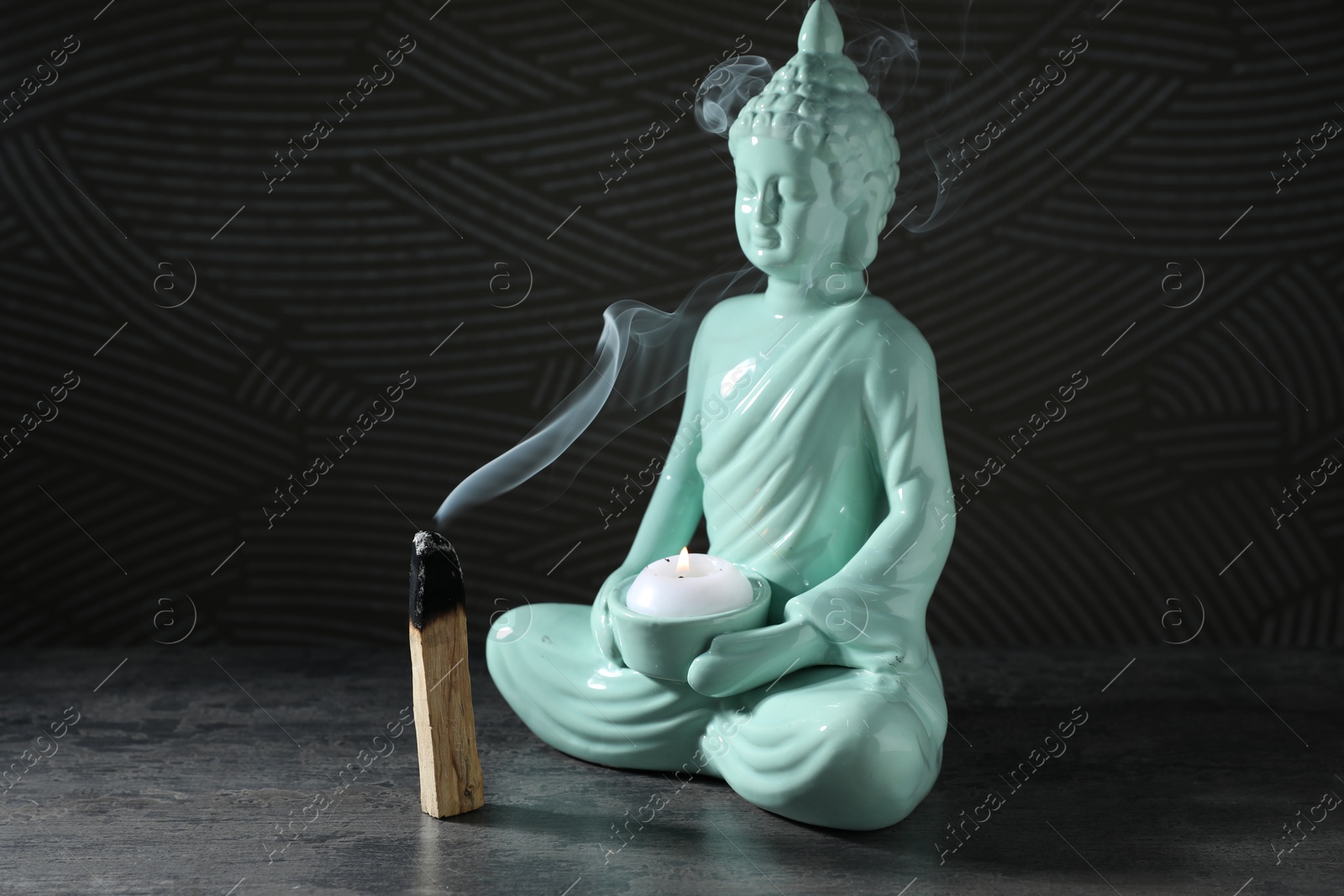 Photo of Smoldering palo santo stick and Buddha statue with burning candle on grey textured table against black background