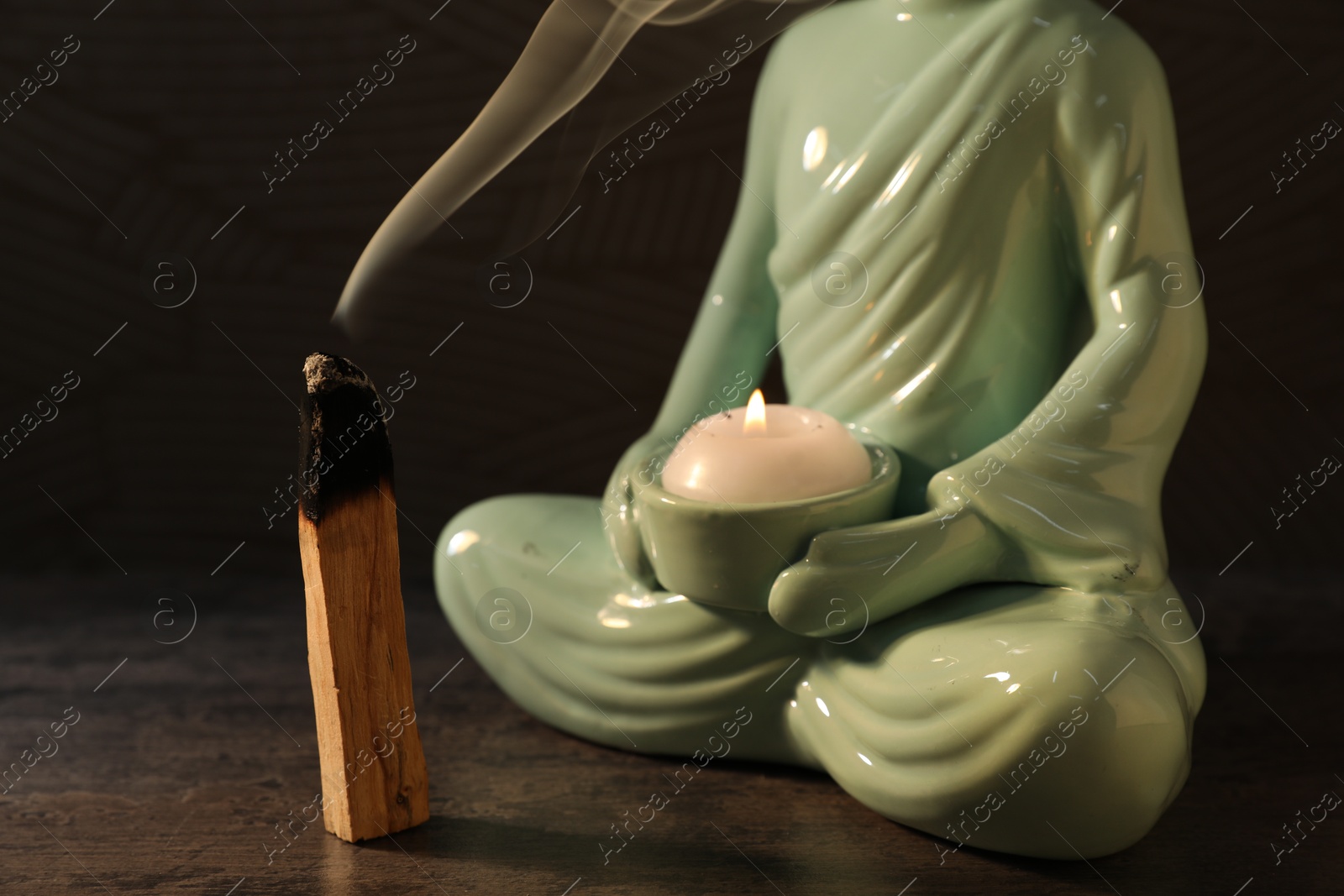 Photo of Smoldering palo santo stick and Buddha statue with burning candle on textured table against dark background, closeup