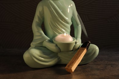 Photo of Smoldering palo santo stick and Buddha statue with burning candle on textured table against dark background, closeup