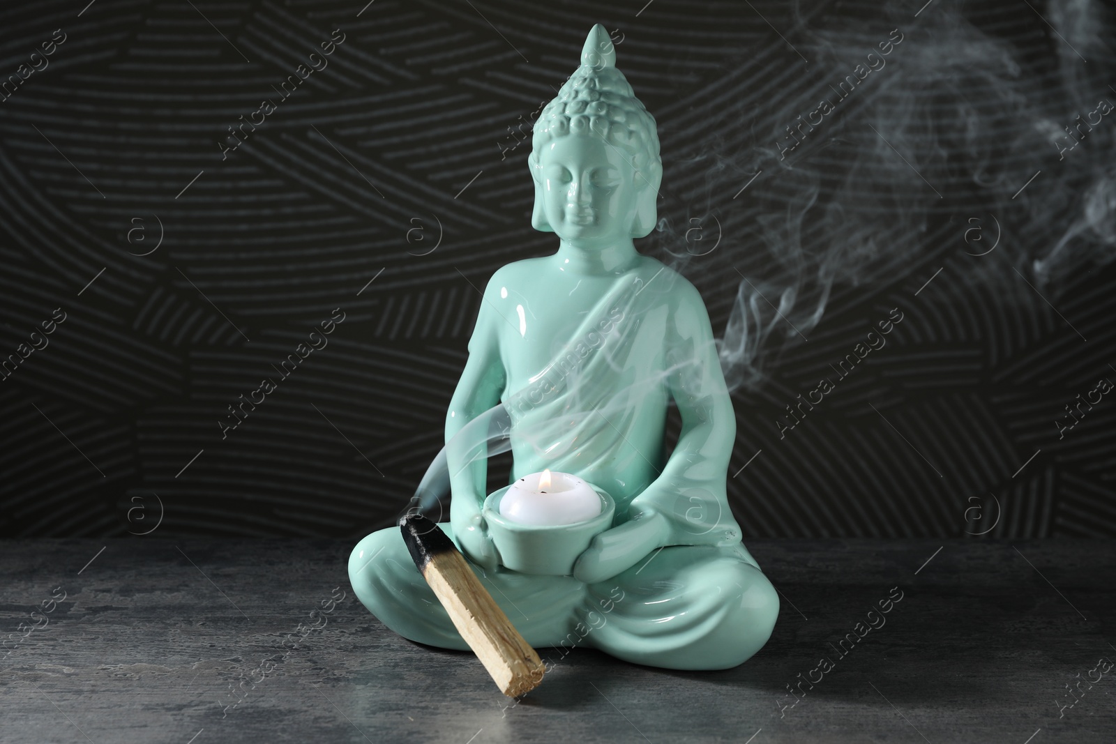 Photo of Smoldering palo santo stick and Buddha statue with burning candle on grey textured table against black background, closeup