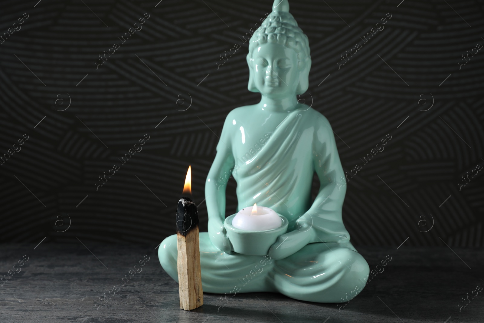 Photo of Burning palo santo stick and Buddha statue with candle on grey textured table against black background, closeup