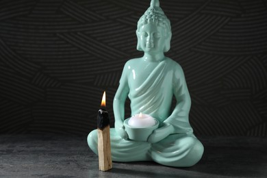 Photo of Burning palo santo stick and Buddha statue with candle on grey textured table against black background, closeup