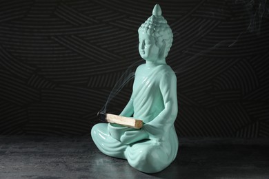 Photo of Smoldering palo santo stick and Buddha statue on grey textured table against black background, closeup