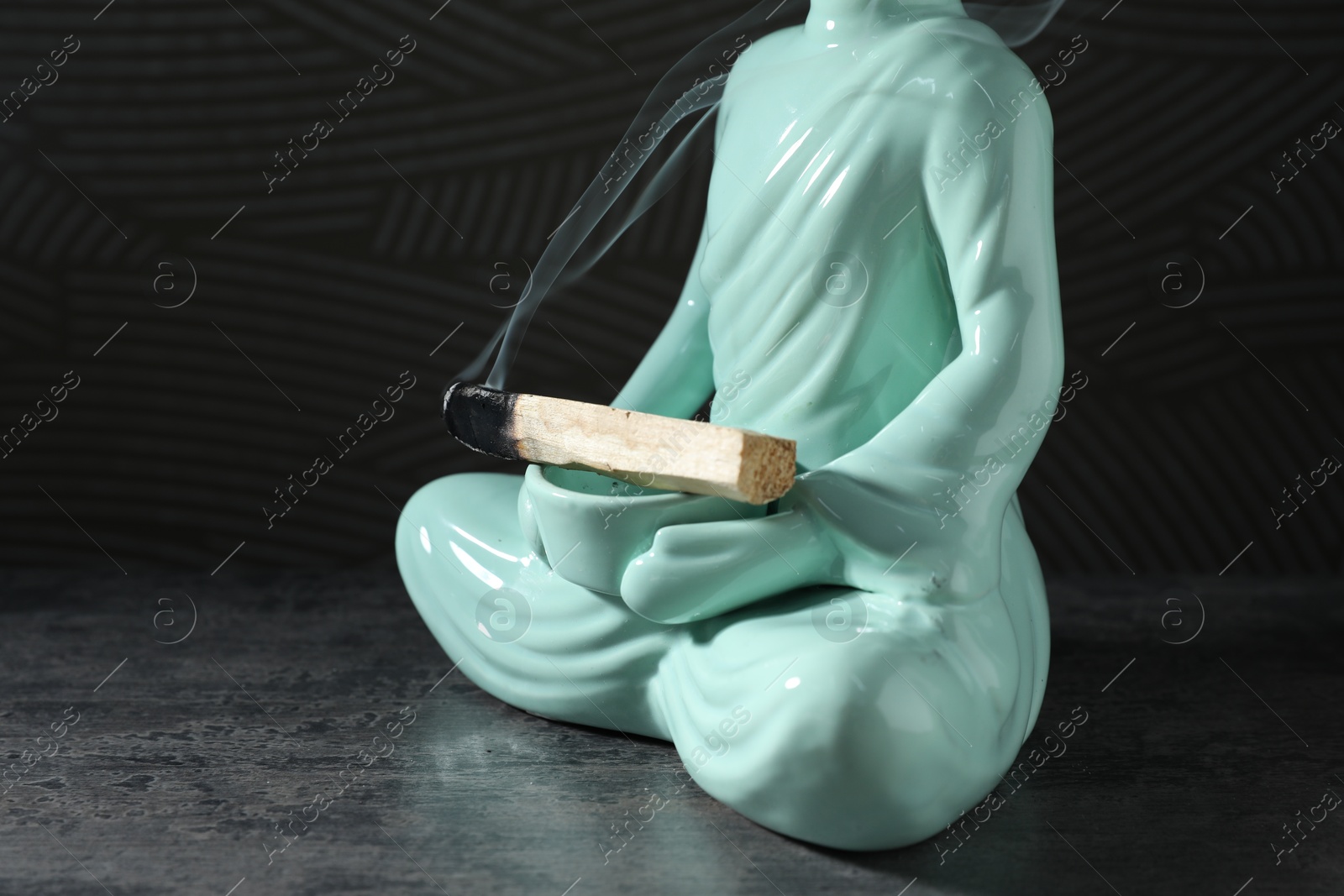 Photo of Smoldering palo santo stick and Buddha statue on grey textured table against black background, closeup