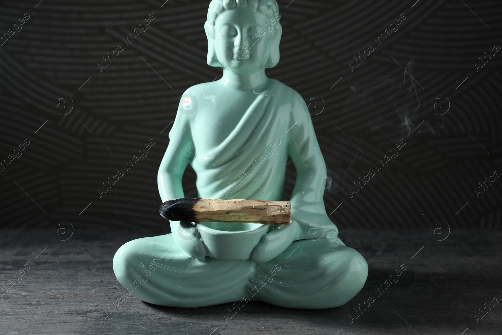 Photo of Smoldering palo santo stick and Buddha statue on grey textured table against black background, closeup