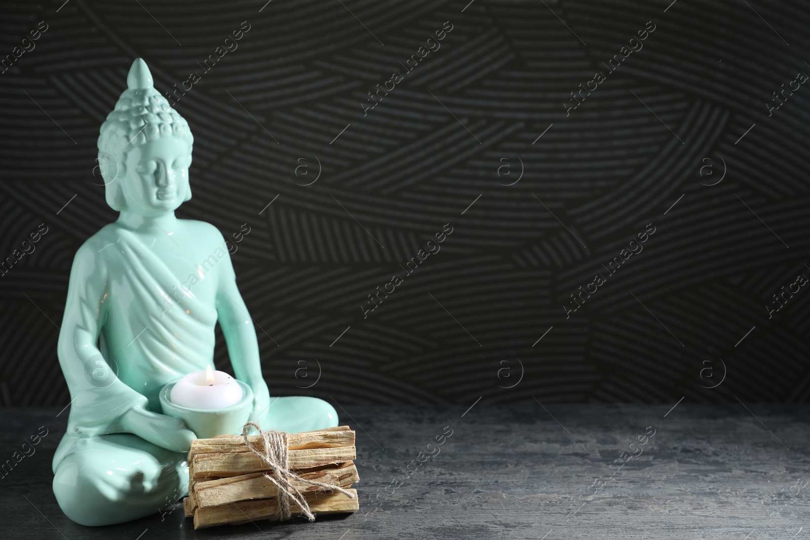 Photo of Palo santo sticks and Buddha statue with burning candle on grey textured table against black background, closeup. Space for text