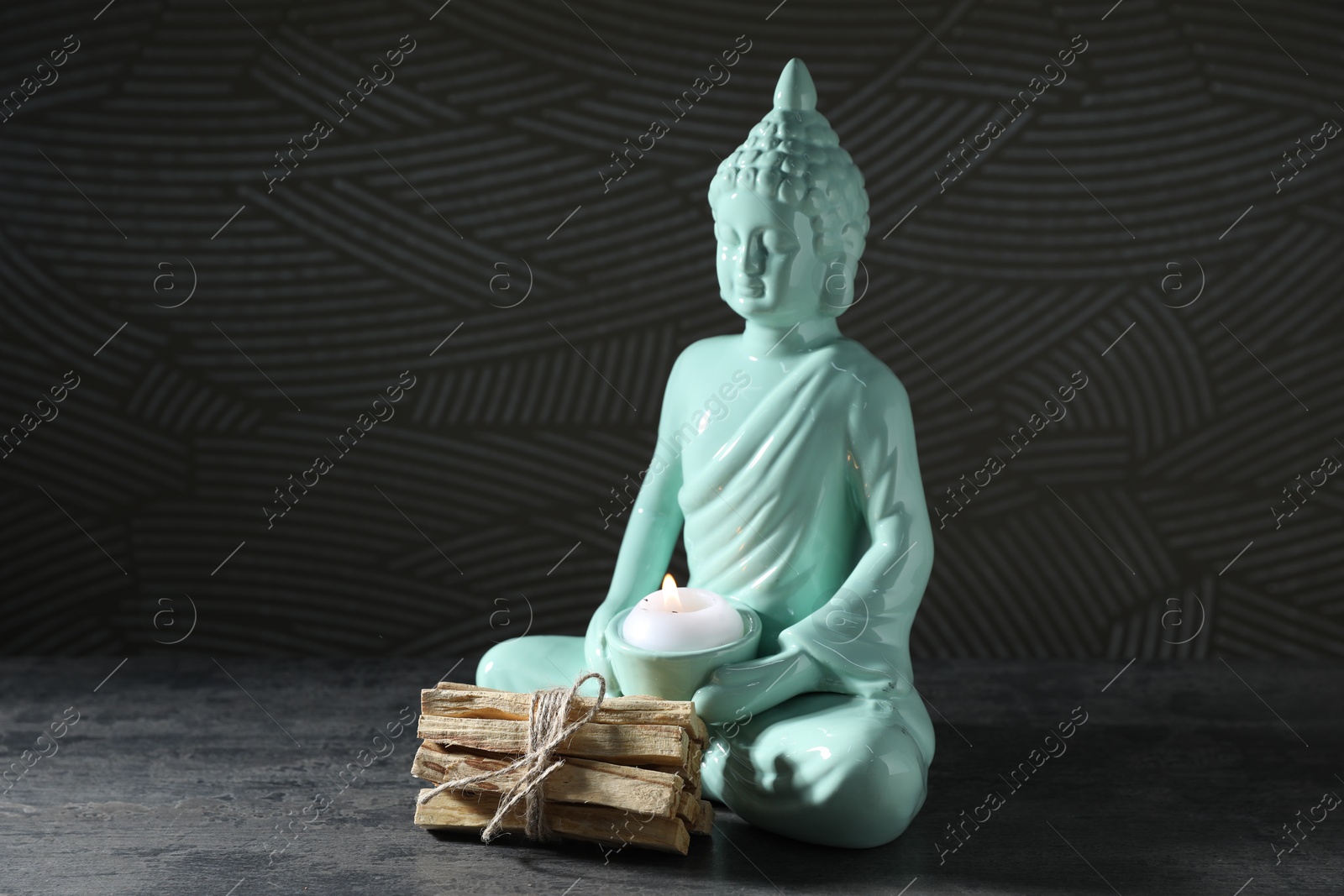 Photo of Bunch of palo santo sticks and Buddha statue with burning candle on grey textured table against black background, closeup