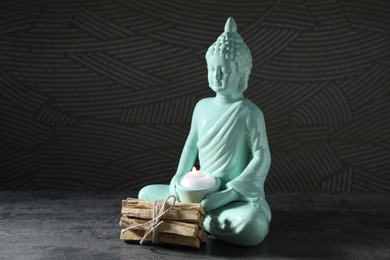 Photo of Bunch of palo santo sticks and Buddha statue with burning candle on grey textured table against black background, closeup