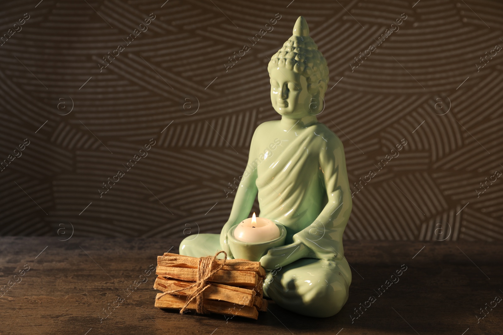 Photo of Bunch of palo santo sticks and Buddha statue with burning candle on grey textured table against black background, closeup