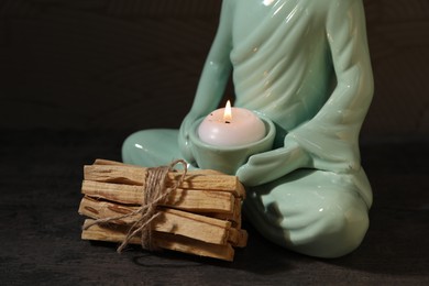 Photo of Bunch of palo santo sticks and statue with burning candle on dark textured table, closeup