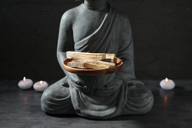 Photo of Bunch of palo santo sticks, statue and burning candles on grey textured table against black background, closeup