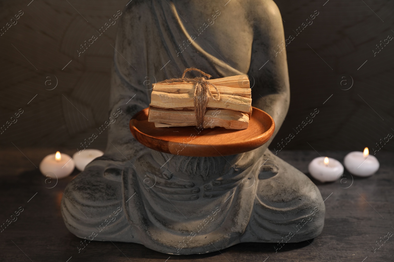 Photo of Bunch of palo santo sticks, statue and burning candles on grey textured table against black background, closeup