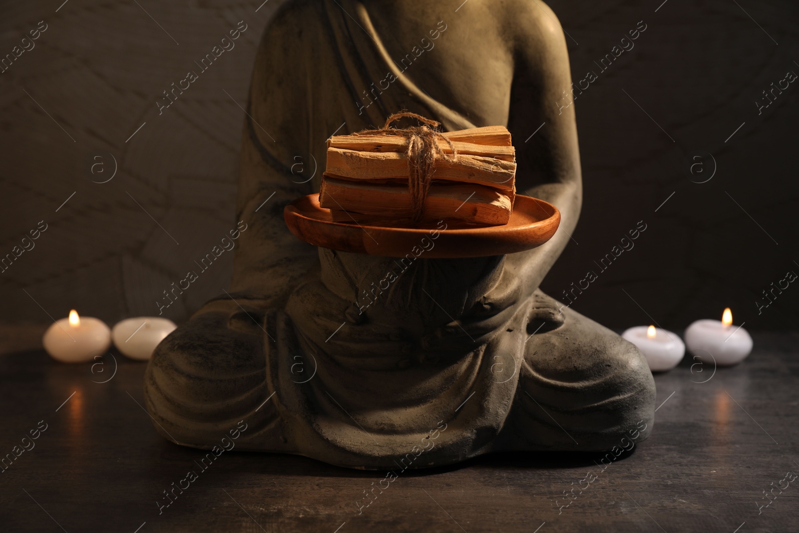 Photo of Bunch of palo santo sticks, statue and burning candles on grey textured table against black background, closeup