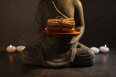 Photo of Bunch of palo santo sticks, statue and burning candles on grey textured table against black background, closeup