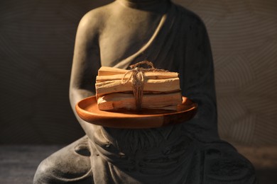 Photo of Bunch of palo santo sticks and statue on table against dark background, closeup