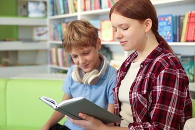 Photo of Teenage friends reading book on sofa in library. Space for text