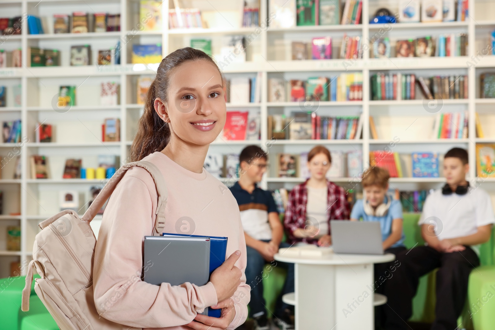 Photo of Portrait of smiling teenage girl with books in public library. Space for text