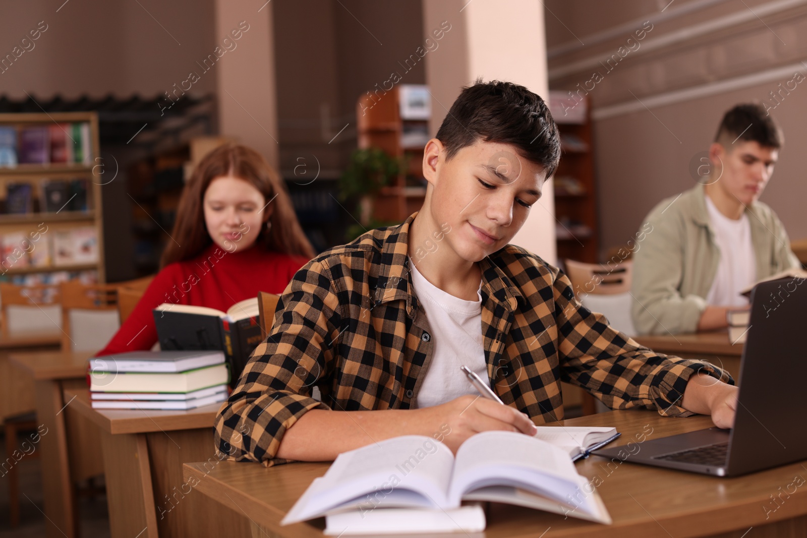 Photo of Boy taking notes while using laptop at desk in public library