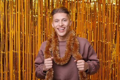 Photo of Happy young man with bright tinsel against golden foil curtain