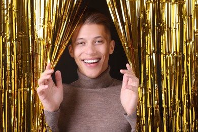 Photo of Happy young man looking out through golden foil curtain against dark background