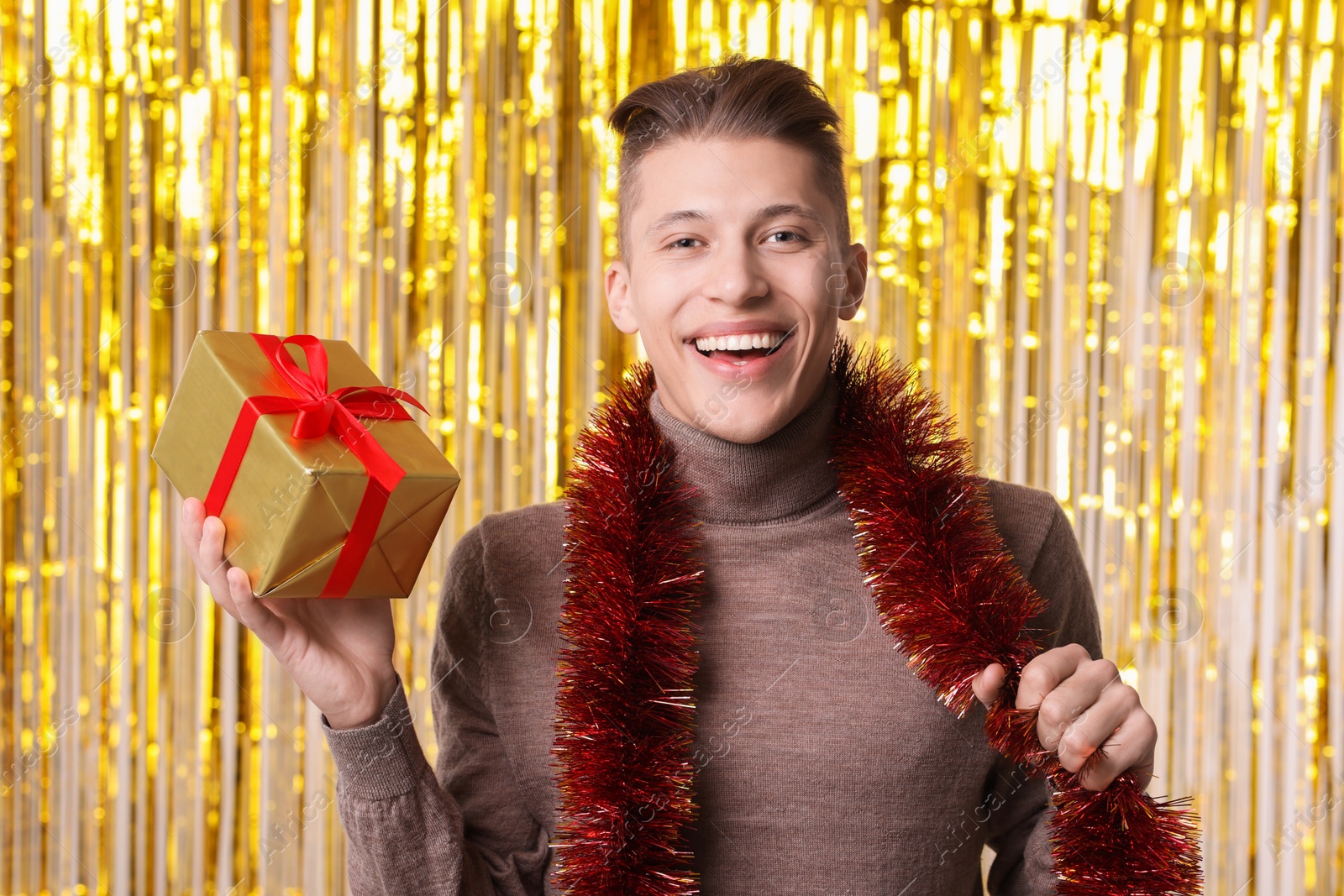 Photo of Happy young man with bright tinsel and gift box against foil curtain