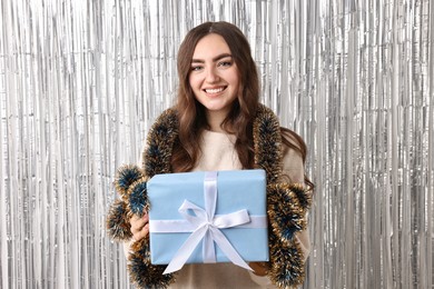 Photo of Happy young woman with tinsel and gift box against silver foil curtain