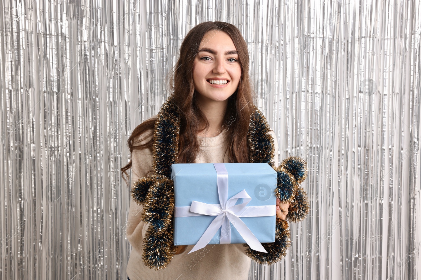 Photo of Happy young woman with tinsel and gift box against silver foil curtain