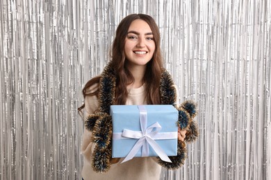 Photo of Happy young woman with tinsel and gift box against silver foil curtain