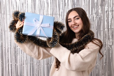 Photo of Happy young woman with tinsel and gift box against silver foil curtain