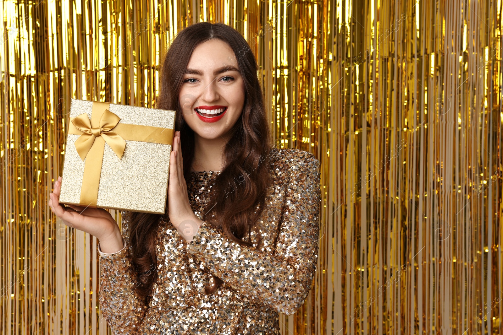 Photo of Happy young woman with gift box against golden foil curtain, space for text