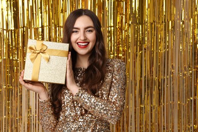 Photo of Happy young woman with gift box against golden foil curtain, space for text