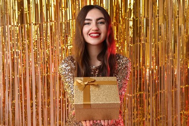 Photo of Happy young woman with gift box against golden foil curtain