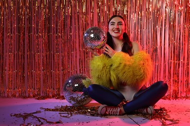 Photo of Beautiful young woman wearing retro outfit with tinsel and disco balls against golden foil curtain in neon lights