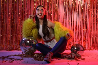 Photo of Beautiful young woman wearing retro outfit with tinsel and disco balls against golden foil curtain in neon lights