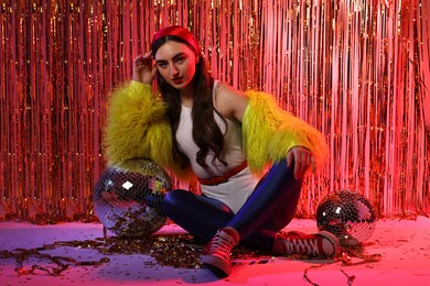 Photo of Beautiful young woman wearing retro outfit with tinsel and disco balls against golden foil curtain in neon lights